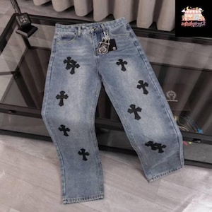 Può includere: Jeans di denim blu chiaro con applicazioni di croci nere. I jeans hanno un classico design a cinque tasche e una vestibilità dritta. Un portachiavi color argento è attaccato al passante per la cintura.