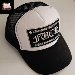 May include: Black and white trucker hat with a mesh back and a curved brim. The front panel is white with a black rectangular patch that reads "CHROME HEARTS FUCK HOLLYWOOD USA" in a gothic font.