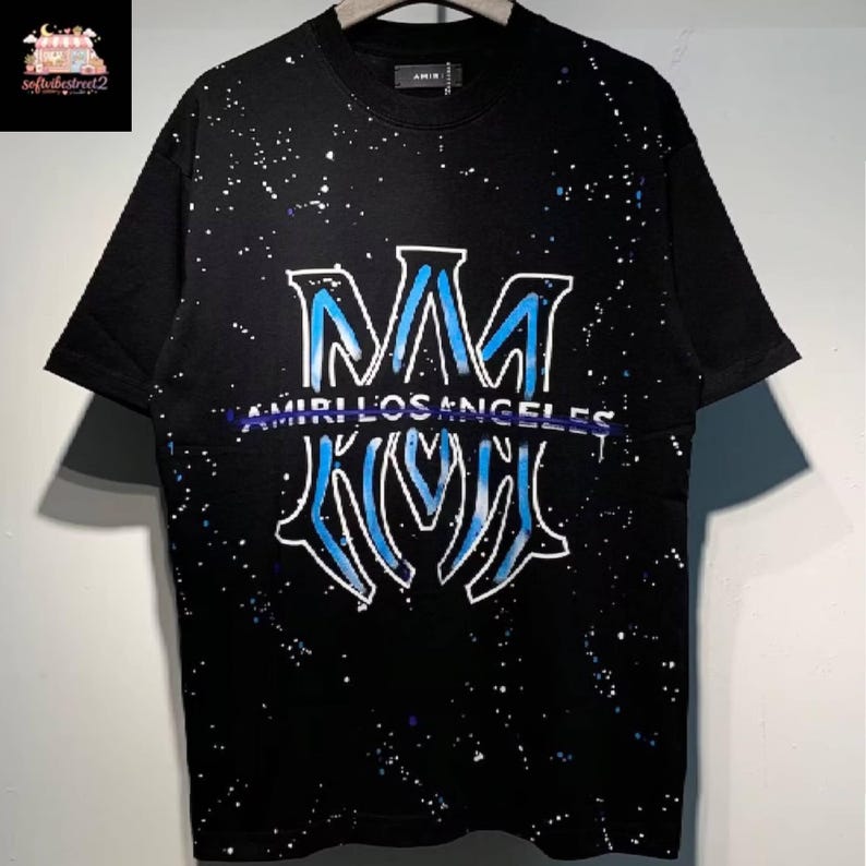 May include: Black t-shirt with a white and blue graphic design. The design features the letters "MM" in a stylized font, with the words "AMIRI LOS ANGELES" underneath. The shirt has a splatter paint effect.
