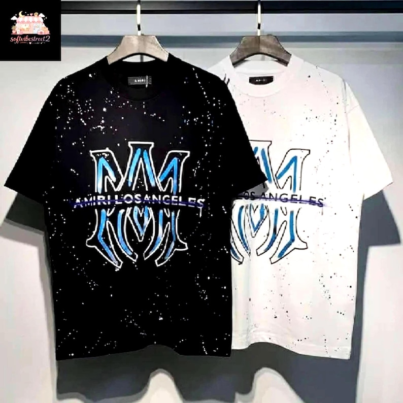 May include: Two short-sleeved t-shirts, one black and one white, are displayed. Both shirts feature a large, stylized "M" design in blue, with the text "AMIRI LOS ANGELES" beneath. The shirts have a splatter paint effect.