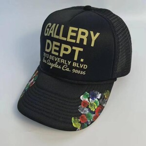 Puede incluir: Gorra de camionero negra con la inscripción dorada "GALLERY DEPT." y la dirección "7613 BEVERLY BLVD Los Angeles Ca. 90036". La visera está decorada con salpicaduras de pintura coloridas.