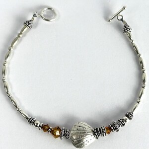 May include: Silver-toned bracelet featuring a heart-shaped charm with a textured surface. The bracelet includes oval beads, decorative accents, and amber-colored crystals. It has a toggle clasp for closure.