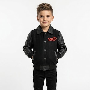 May include: A black varsity jacket featuring black leather sleeves, a black wool-blend body, a pointed collar, and a front button closure. The word BAD is embroidered in red on the upper left chest. The jacket has two side pockets and ribbed cuffs.