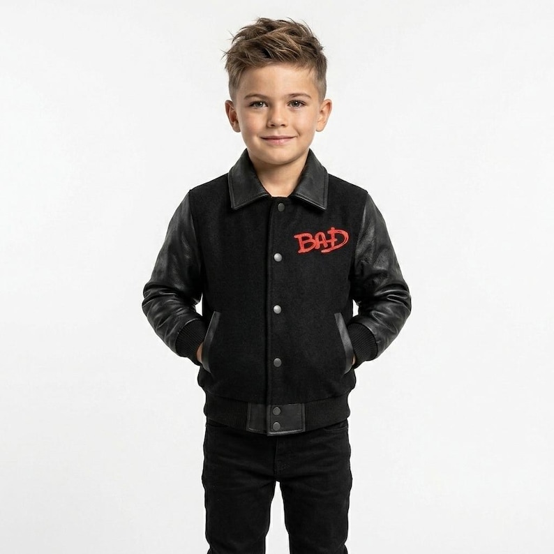 May include: A black varsity jacket featuring black leather sleeves, a black wool-blend body, a pointed collar, and a front button closure. The word BAD is embroidered in red on the upper left chest. The jacket has two side pockets and ribbed cuffs.
