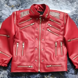 May include: A bright red jacket featuring a central silver zipper, multiple horizontal and vertical silver zippered pockets, and silver stud accents. The shoulders are adorned with silver metallic panels, and the sleeves have elastic cuffs.