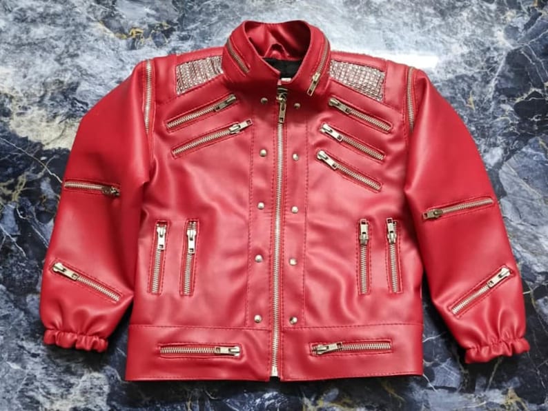 May include: A bright red jacket featuring a central silver zipper, multiple horizontal and vertical silver zippered pockets, and silver stud accents. The shoulders are adorned with silver metallic panels, and the sleeves have elastic cuffs.