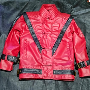 May include: A bright red jacket featuring black leather-like accents, a high collar with ribbed detailing, and a front closure with five large red buttons. The garment includes black straps with gold-toned buckles on the cuffs and waist.