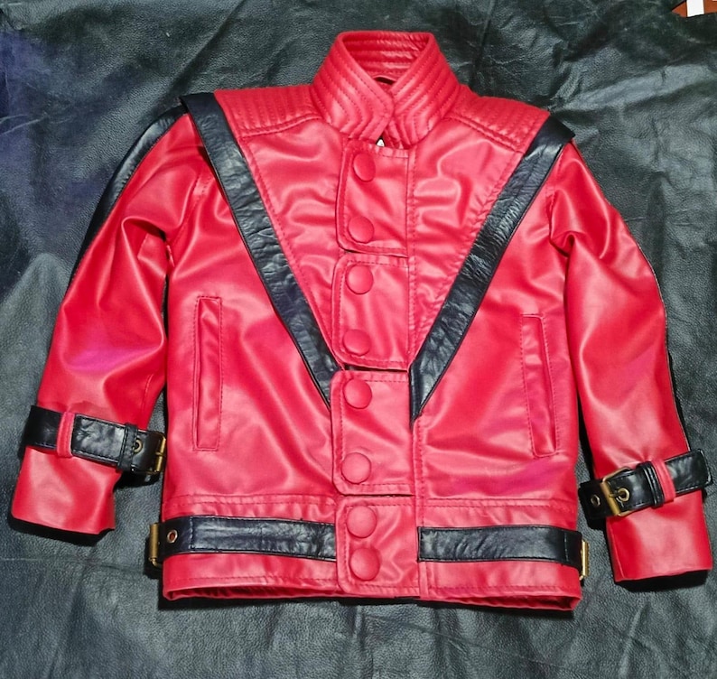 May include: A bright red jacket featuring black leather-like accents, a high collar with ribbed detailing, and a front closure with five large red buttons. The garment includes black straps with gold-toned buckles on the cuffs and waist.