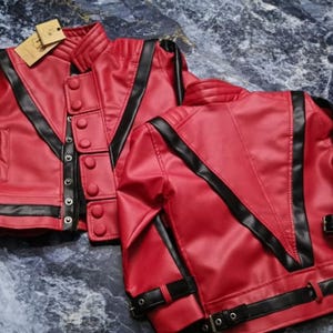 May include: A bright red jacket with black trim, featuring a high collar, a front button closure, and black buckled straps on the sleeves and hem. The jacket has a V-shaped black panel design on the front and back, with a small tan tag attached.