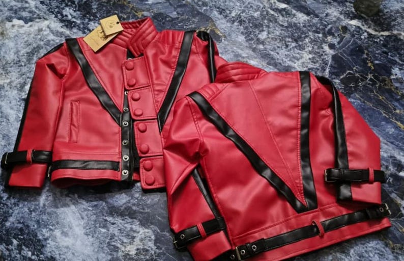 May include: A bright red jacket with black trim, featuring a high collar, a front button closure, and black buckled straps on the sleeves and hem. The jacket has a V-shaped black panel design on the front and back, with a small tan tag attached.