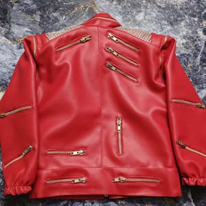 May include: A bright red jacket featuring multiple gold-toned metal zippers across the front, sleeves, and chest. The shoulders are embellished with a dense pattern of small, reflective silver studs, and the cuffs are gathered with elastic.