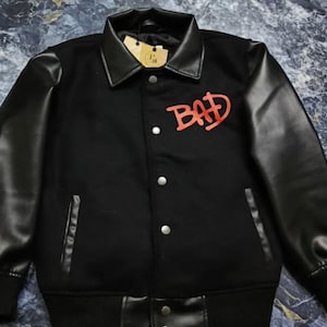 May include: A black varsity-style jacket featuring long black sleeves, a pointed collar, and a front button closure. The chest displays the word BAD in red lettering. The garment includes two side pockets and a ribbed hem and cuffs.
