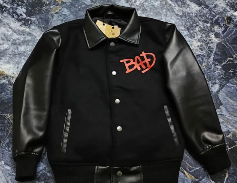 May include: A black varsity-style jacket featuring long black sleeves, a pointed collar, and a front button closure. The chest displays the word BAD in red lettering. The garment includes two side pockets and a ribbed hem and cuffs.