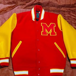 May include: A red varsity jacket with yellow sleeves, featuring a large yellow letter M on the left chest. The jacket has a snap-button front closure, a ribbed collar with white stripes, and ribbed cuffs and hem with white and red striped detailing.