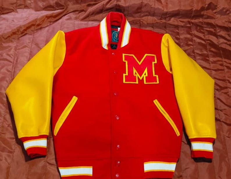 May include: A red varsity jacket with yellow sleeves, featuring a large yellow letter M on the left chest. The jacket has a snap-button front closure, a ribbed collar with white stripes, and ribbed cuffs and hem with white and red striped detailing.
