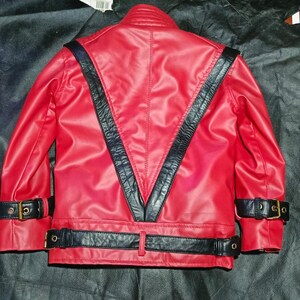 May include: A bright red jacket featuring a prominent black V-shaped panel on the back. The garment includes black buckled straps at the cuffs and a matching black buckled belt around the waist, all constructed from a smooth, leather-like material.