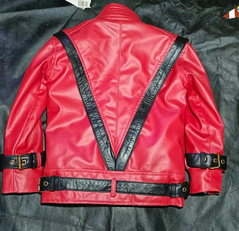 May include: A bright red jacket featuring a prominent black V-shaped panel on the back. The garment includes black buckled straps at the cuffs and a matching black buckled belt around the waist, all constructed from a smooth, leather-like material.