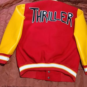 May include: A red varsity jacket with bright yellow sleeves. The back features the word THRILLER in large white letters with black outlines. The jacket has a ribbed collar, cuffs, and hem with white and yellow stripes, and two visible snap buttons.