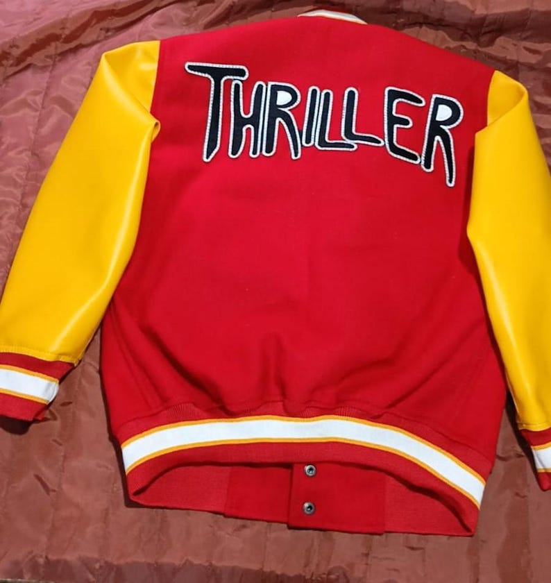 May include: A red varsity jacket with bright yellow sleeves. The back features the word THRILLER in large white letters with black outlines. The jacket has a ribbed collar, cuffs, and hem with white and yellow stripes, and two visible snap buttons.