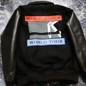 May include: A black varsity-style jacket with long black sleeves. The back features a graphic print with the text MICHAEL JACKSON in a red rectangle, a white silhouette of a dancer, and the text WORLD TOUR in a blue rectangle below.
