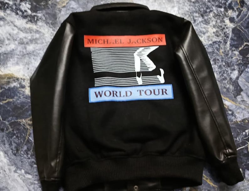 May include: A black varsity-style jacket with long black sleeves. The back features a graphic print with the text MICHAEL JACKSON in a red rectangle, a white silhouette of a dancer, and the text WORLD TOUR in a blue rectangle below.