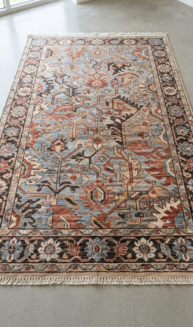 May include: A rectangular area rug featuring a complex pattern of floral and geometric motifs in shades of light blue, rust orange, dark brown, and cream. The rug has a wide decorative border and short fringe along the top and bottom edges.