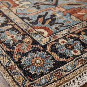May include: A rectangular area rug featuring a complex pattern of floral and geometric motifs in shades of light blue, rust orange, dark navy, and beige. The rug has a thick, textured pile and is finished with short, off-white fringe at the ends.