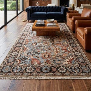 May include: A rectangular area rug featuring a complex floral and geometric pattern in shades of blue, rust, cream, and black. The rug has a dark border with repeating flower motifs and short, light-colored fringe along the short edges.
