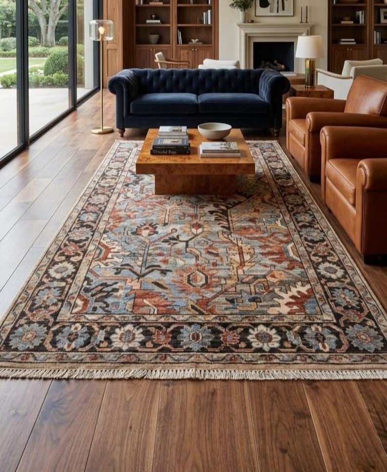 May include: A rectangular area rug featuring a complex floral and geometric pattern in shades of blue, rust, cream, and black. The rug has a dark border with repeating flower motifs and short, light-colored fringe along the short edges.