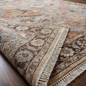 May include: A rectangular area rug featuring a complex floral and geometric pattern in shades of dusty blue, terracotta, and cream. The rug has a dark brown border with intricate detailing and a short, cream-colored fringe along the edge.