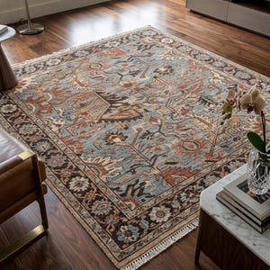 May include: A rectangular area rug featuring a complex pattern in shades of dusty blue, terracotta, cream, and dark brown. The rug has a wide decorative border with floral motifs and short white fringe along the edges, laid flat on a dark wood floor.