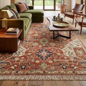 Op de afbeelding: Een groot rechthoekig vloerkleed met een terracotta rode basis en ingewikkelde bloem- en geometrische patronen in marineblauw, crème en saliegroen. Het kleed heeft decoratieve franjes aan de korte zijden en een geweven textuur.