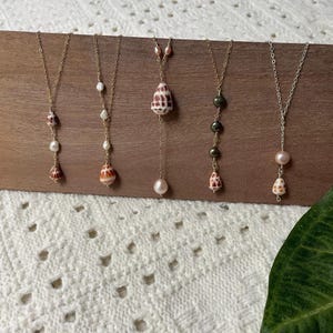 Op de afbeelding: Vijf goudkleurige kettingen gepresenteerd op een houten plank. Elke ketting heeft een fijne schakelketting versierd met diverse elementen, waaronder witte parels, donkere kralen en kleine schelpen met bruine en crèmekleurige patronen.
