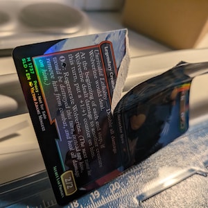 May include: A holographic trading card featuring the text Unrivaled Lethality and Wolverine, partially torn to reveal the white paper core. The card displays game mechanics and the artist name Victor Adame Minguiz, positioned over a ruler.