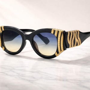 Puede incluir: Gafas de sol negras con detalles decorativos dorados. Las gafas tienen una lente degradada que pasa de amarillo a azul. Las monturas son gruesas y presentan un diseño audaz y geométrico.
