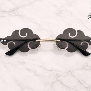 May include: Black cloud-shaped sunglasses with a gold bridge and black arms. The sunglasses have a unique design, with the lenses cut out in the shape of clouds. The frame has two small white dots on each side.