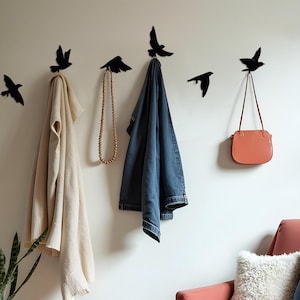 Op de afbeelding: Woonkamer met een koraalrode fauteuil, kussens en een raam. Zwarte vogelvormige wandhaken houden een jas, een handtas en een ketting vast. Een geweven mand bevat een hoge plant.