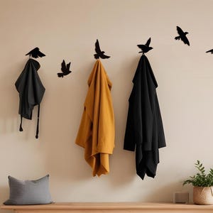 May include: A wall-mounted coat rack featuring black bird silhouettes. Three coats in black and mustard yellow hang from the hooks. A gray pillow rests on a wooden bench below. A small potted plant sits to the right.