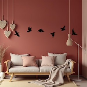 Op de afbeelding: Een woonkamerscène met een houten bank, kussens en een deken. Hartvormige decoraties hangen aan het plafond. Zwarte vogel silhouetten sieren de rode muur, en een vloerlamp staat in de buurt. Een vaas met gedroogde planten staat links.