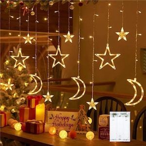 May include: String lights with star and crescent moon shapes. The lights are warm white and hang from a dark background. A wooden sign that says "Happy Holidays" is visible, along with wrapped gifts and decorative lights.