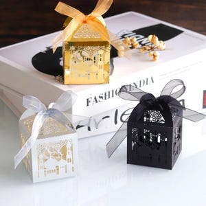 Puede incluir: Tres cajas de regalo decorativas en oro, blanco y negro, cada una con un diseño de mezquita cortado con láser. Las cajas están atadas con cintas transparentes en colores a juego. La caja dorada tiene una cinta naranja, la caja blanca tiene una cinta plateada y la caja negra tiene una cinta negra.