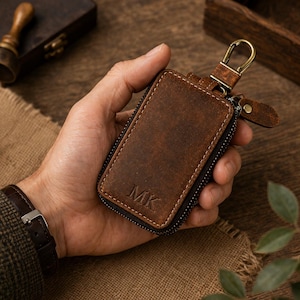 Könnte beinhalten: Ein brauner Leder-Schlüsselhalter mit Reißverschluss und Metallverschluss. Die Initialen "MK" sind auf der Vorderseite eingeprägt. Der Schlüsselhalter wird in einer Hand gehalten, um seine Größe und Textur zu zeigen.