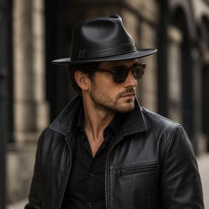 May include: A black fedora hat with a matching black band, worn with a black leather jacket, a black collared shirt, and brown-rimmed sunglasses. The hat features a structured crown and a wide brim, providing a classic look with clean lines.