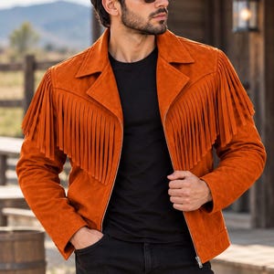 May include: A burnt orange suede jacket featuring long fringe detailing across the chest and back, a classic collar, and a full-length front zipper closure. The jacket is worn over a plain black t-shirt and paired with black denim jeans.