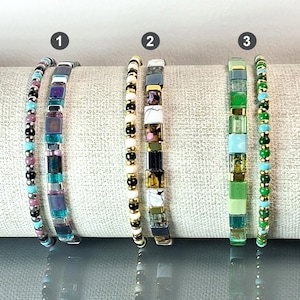 Peut inclure: Trois bracelets de perles aux designs variés. Le bracelet 1 présente des perles violettes, turquoise et noires. Le bracelet 2 comporte des perles dorées, noires et blanches. Le bracelet 3 comprend des perles vertes, bleues et dorées. Tous les bracelets sont présentés sur une surface neutre.