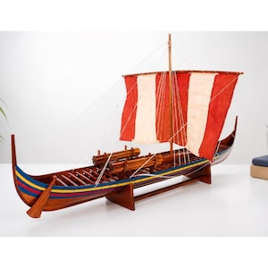 Puede incluir: Un modelo de barco de madera con un casco decorado con franjas pintadas en azul, amarillo y rojo. La embarcación tiene un mástil con una vela rectangular dividida en paneles verticales rojos y blancos, sujeta por finos cabos y remos.