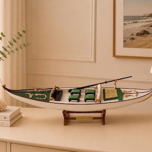 May include: A decorative model gondola featuring a white hull with gold accents, green interior seating, and a black oar. The boat rests on a brown wooden stand with a small plaque that reads GONDOLA in gold lettering, showcasing intricate details.