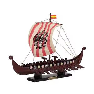 May include: A wooden Viking ship model with a red and white striped sail featuring a crest. The ship has a dark brown hull, oars, and a small flag. The model is displayed on a black base.