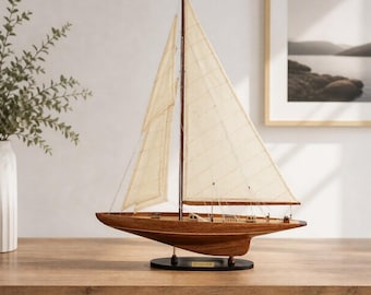 May include: A wooden model sailboat featuring a polished brown hull, two off-white fabric sails, and thin rigging lines. The vessel is mounted on a black oval base with a small gold-colored plaque attached to the center of the stand.