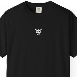 May include: A black short-sleeve t-shirt featuring a small white graphic of a stylized bull head logo centered on the chest. The shirt has a crew neckline and a visible brand label printed on the inside of the collar area at the back neck.